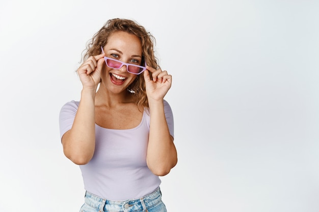 Attractive young coquettish girl looks in sunglasses, smiles happy, stands white. Copy space