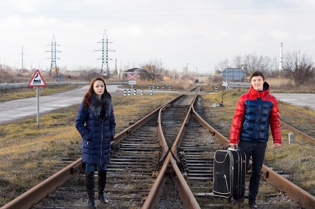 Attractive young couple in fashionable warm clothing standing waiting on a railroad track for the train to arrive with a packed suitcase