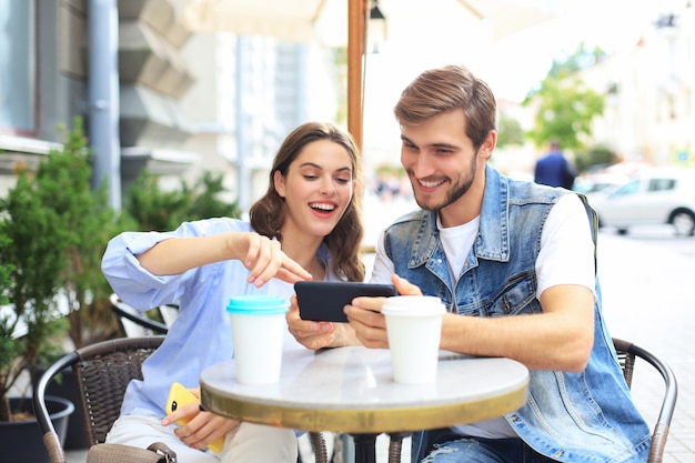 Attractive young couple in love drinking coffee while sitting at the cafe table outdoors, using mobile phone.