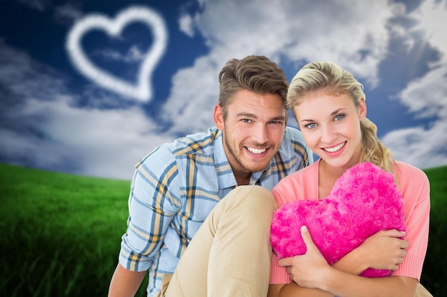 Attractive young couple sitting holding heart cushion against green field under blue sky