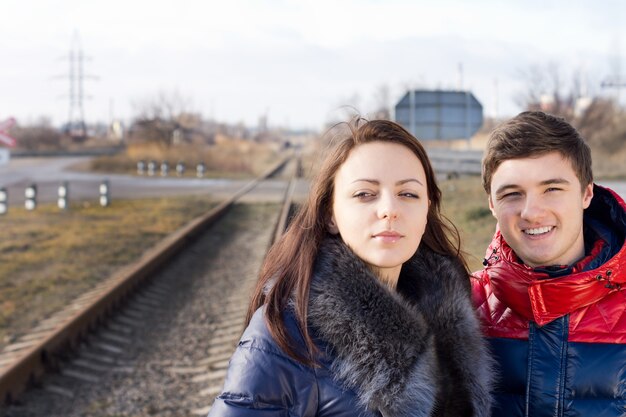 電車が到着するのを線路の横で待っている一緒に立っている暖かいキルティングの服を着た魅力的な若いカップル