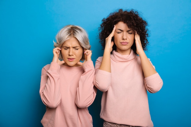 Attractive young curly woman with her female best friend touching their head with both because having headache isolated on blue wall.