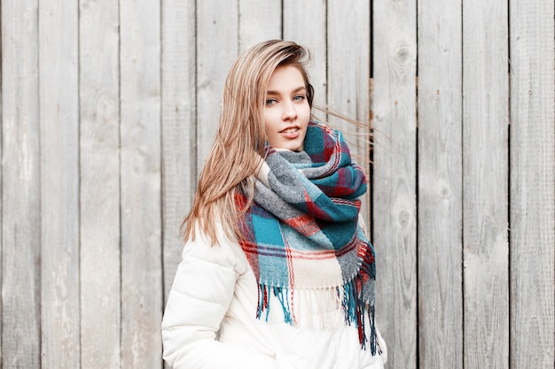 Attractive young cute woman in a stylish white warm jacket with a woolen fashionable warm colorful scarf standing on the street near the wooden vintage wall. Girl with beautiful blue eyes