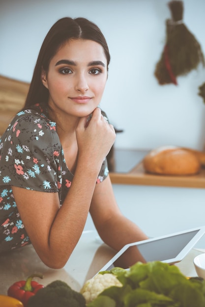 An attractive young darkhaired woman chooses the recipe for a delicious meal while sitting at the table in the kitchen Tablet pc is the best cookbook