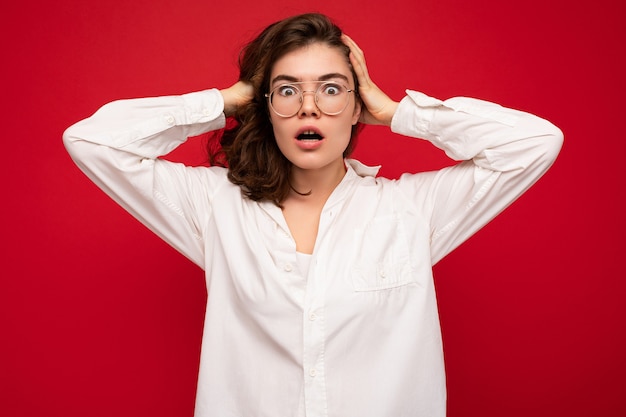 Attractive young emotional shocked amazed curly brunette woman wearing white shirt and optical