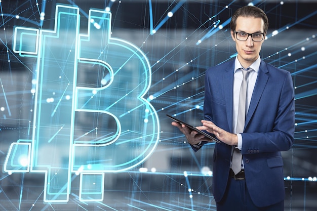 Attractive young european businessman using tablet with abstract glowing holographic bitcoin icon on blurry office interior background Cryptocurrency online banking and money concept Double exposure