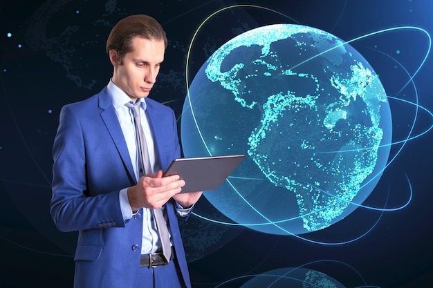 Attractive young european businessman using tablet with creative glowing globe hologram on dark background Technology metaverse and digital map concept