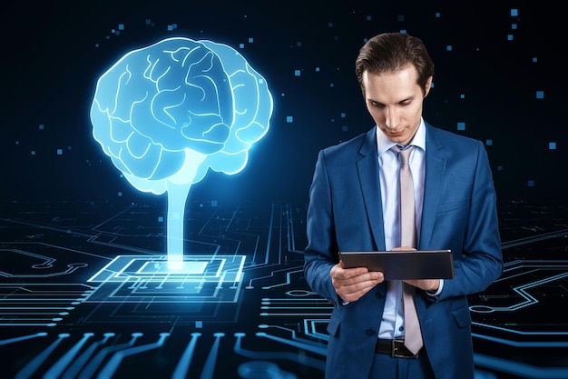 Attractive young european businessman using tablet with glowing circuit brain hologram on dark background AI machine learning and futuristic technology concept