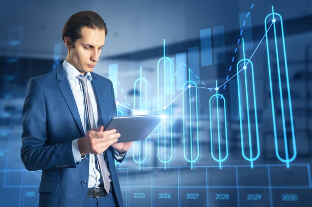 Attractive young european businessman using tablet with growing annual business chart report hologram on blurry office interior background Analysis and company concept Double exposure