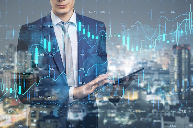 Attractive young european businessman with pad standing on\
abstract double exposure city background with forex chart and graph\
finance stock market and communication concept