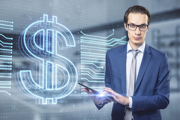 Attractive young european businessman with tablet glowing dollar hologram on blurry office interior background Money banking app and finance concept Double exposure