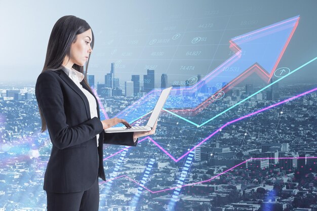 Attractive young european businesswoman using laptop with\
abstract growing business arrow and chart on blurry city background\
with index grid financial data trade and market concept double\
exposure