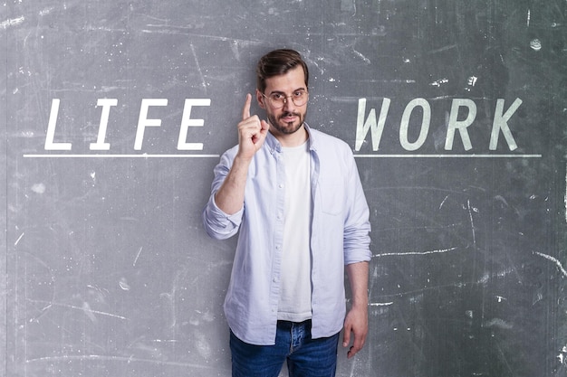 Attractive young european man in glasses and with writing on\
concrete wall pointing up and trying to balance life and work\
success career failure work life and personal development\
concept