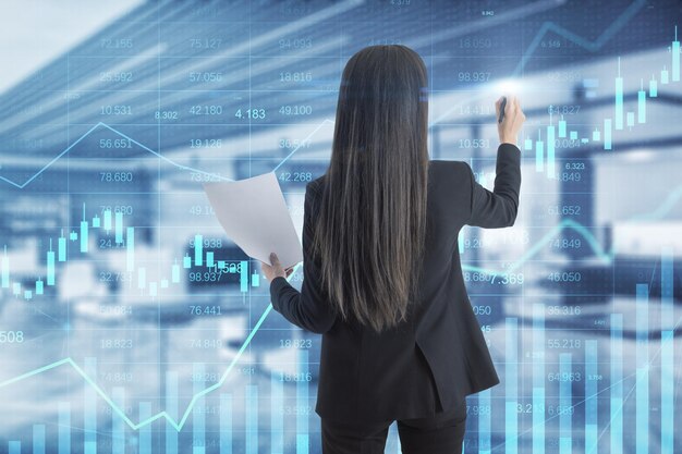 Attractive young european woman using glowing business chart\
hologram and index on blurry office interior background finance\
trade and invest concept double exposure