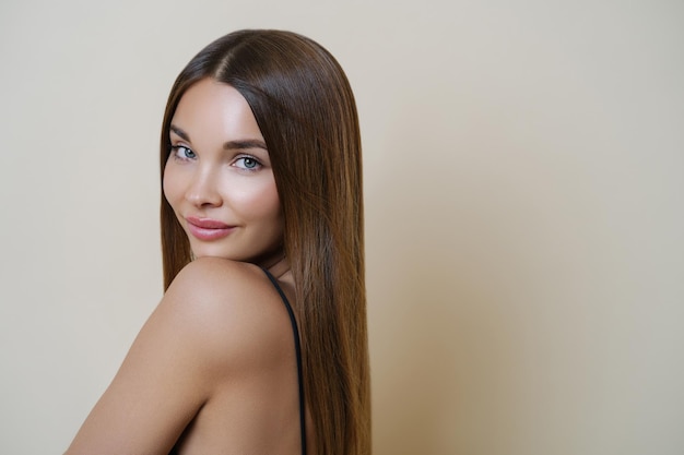 Attractive young European woman with soft healthy skin perfect body looks calm directly at camera has long hair stands indoor against beige background Youth beauty and skin care concept