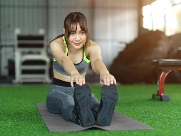 Attractive young female doing warmup stretching exercise before workout, sits on exercise mat in sport centre.