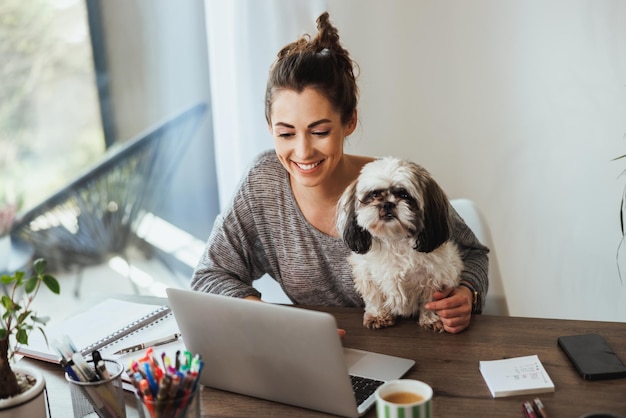 Attractive young female freelancer working on laptop from her home and having her pet dog in her lap to keep her company.