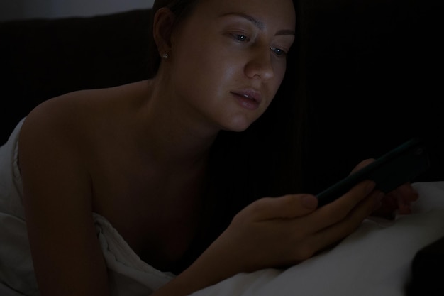 Attractive young female lying in comfortable bed late at night and using mobile phone before sleep