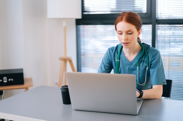 attractive young female practitioner doctor in blue green medical uniform working typing on laptop 