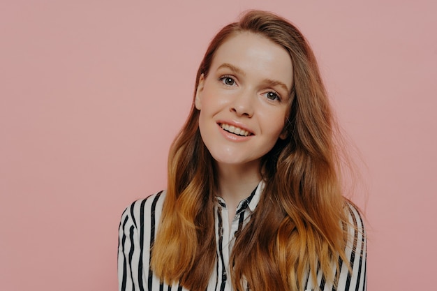 Attractive young female with wavy medium length ginger hair in stripy black and white blouse tilting head while posing over light pink background. Human emotions concept