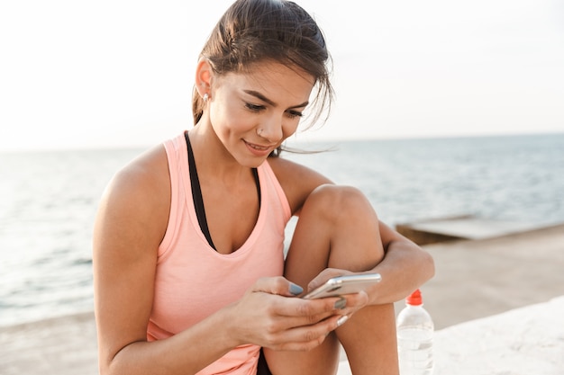 Attractive young fitness woman using mobile phone while resting at the beach after jogging