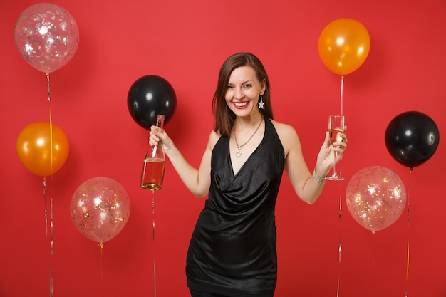 Attractive young girl in black dress celebrating hold glass bottle of champagne on bright red background air balloons. International Women's Day, Happy New Year, birthday mockup holiday party concept.