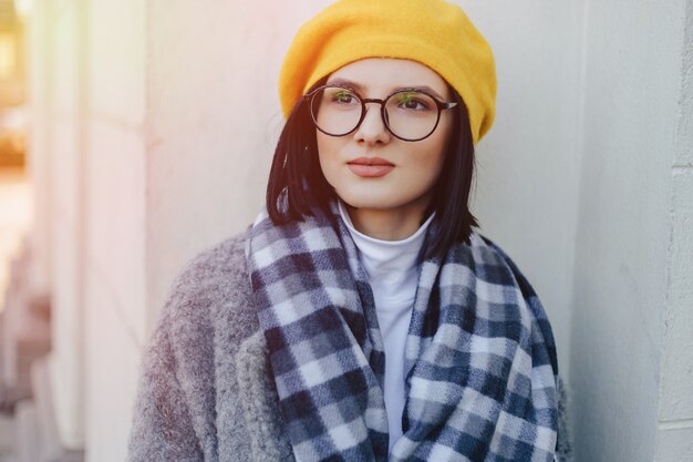 Attractive young girl in glasses in coat and yellow Beret on a simple light background