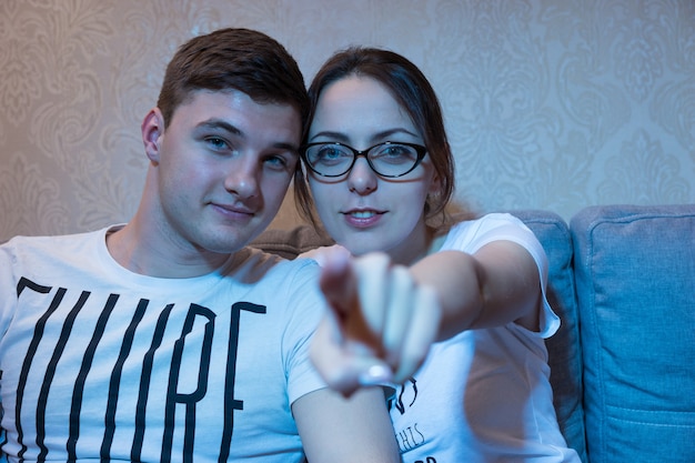 La ragazza attraente sta indicando la macchina fotografica che si siede su un divano con il suo ragazzo a casa guardando la televisione, vista frontale nel bagliore blu dal set