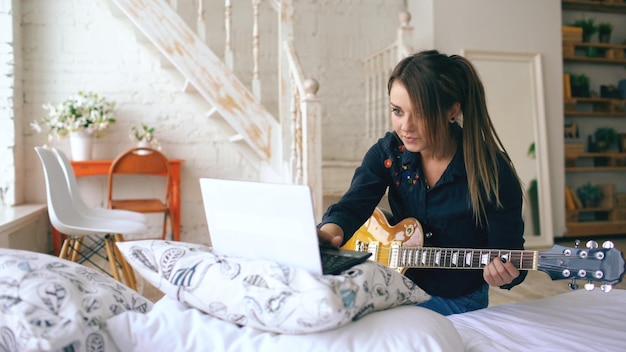 Attractive young girl learning to play electric guitar with notebook sit on bed in bedroom at home