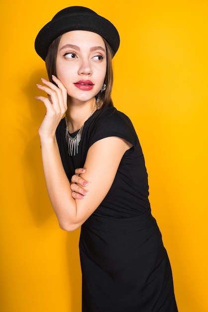 Attractive young girl model in black dress and hat posing, in expensive jewelry, on yellow background
