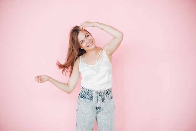 Attractive young girl in summer clothes gave a lot of fun and plays with her hair, picture isolated on pink background