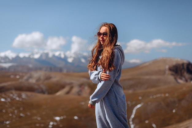 An attractive young girl in a warm blue jumpsuit and sunglasses stands and looks at the mountain landscape