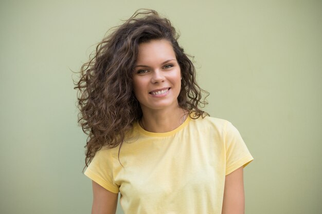 Attractive young girl with brown curly hair standing in a yellow t-shirt against a green wall