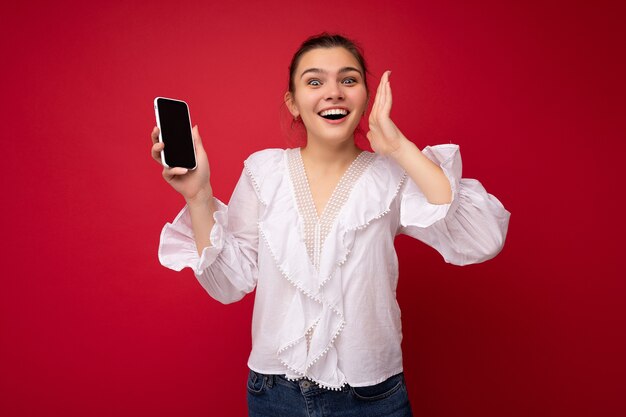 赤い背景の上に孤立して立っている白いブラウスを着て魅力的な若い幸せな黒髪の女性