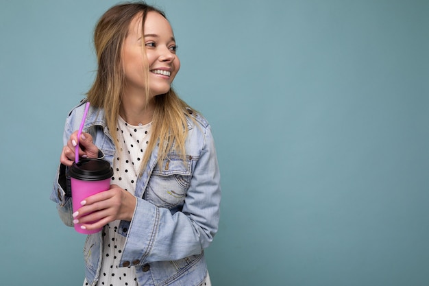 Attractive young happy smiling blonde woman wearing everyday stylish clothes isolated over colourful background wall holding paper cup for cutout drinking tea looking to the side. copy space