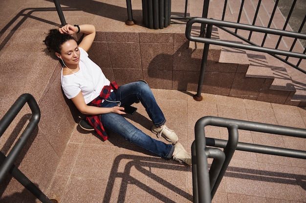Attractive young Hispanic woman listening to music on headphones and having rest leaning on steps of unrecognizable building, enjoying warm sunny day