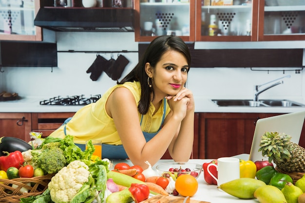 Attractive young Indian girl with apron cooking in kitchen with table full of fruits and vegetables