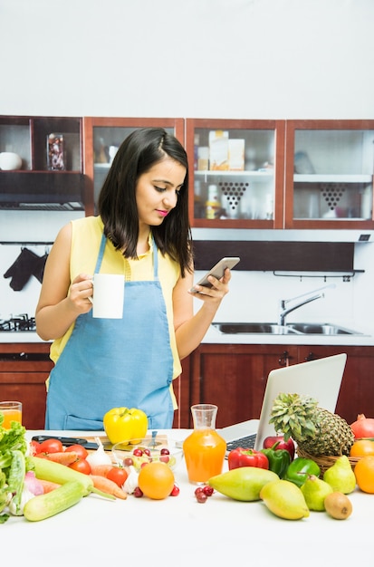 Привлекательная молодая индийская девушка или женщина с фартуком на кухне многозадачности, используя смартфон, планшетный ПК со столом, полным фруктов и овощей и компьютером