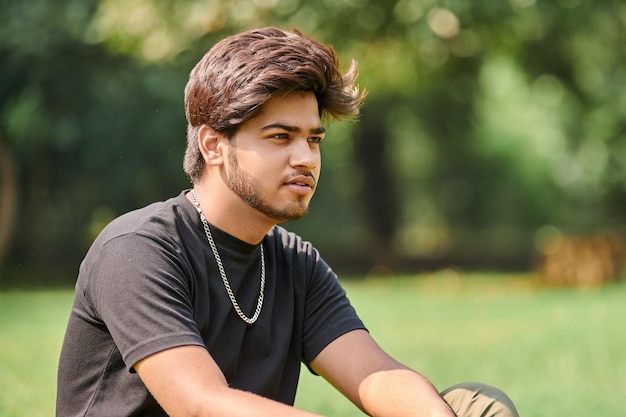 Attractive young indian man portrait in black t shirt and silver neck chain sitting on green lawn