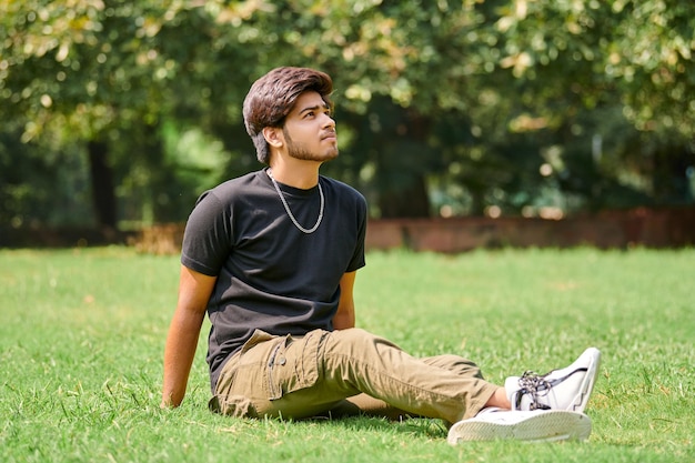 Attractive young indian man portrait in black t shirt and silver neck chain sitting on green lawn