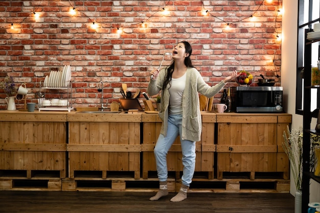 Attractive young joyful asian woman have fun alone dancing and\
singing while cook in kitchen at home. female in winter wear socks\
relax enjoy free time in house red brick wall in back light\
hanging