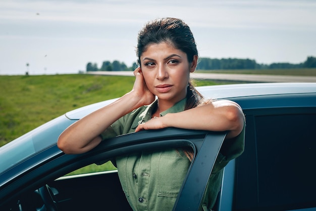 Attraente giovane donna è appoggiata alla portiera della sua auto e guarda in lontananza, ritratto