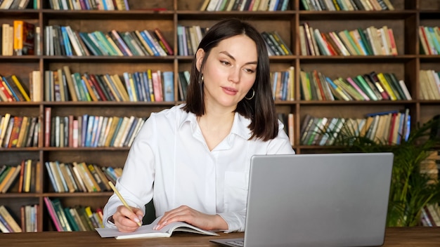 Attractive young lady in stylish white blouse writes in paper notebook at videocall on contemporary laptop at table against racks of books