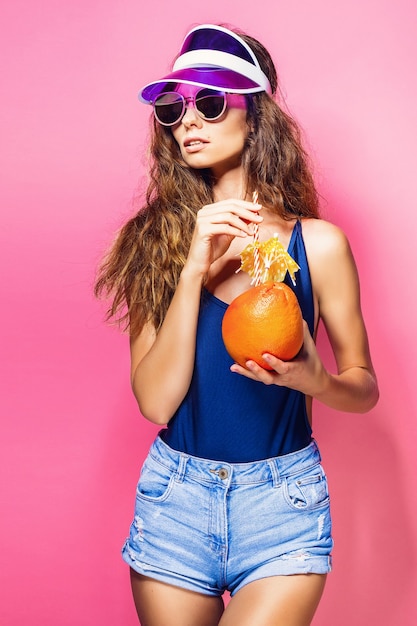 Attractive young lady in summer outfit holding fresh juicy citrus with straw and umbrella and looking at camera while standing on pink