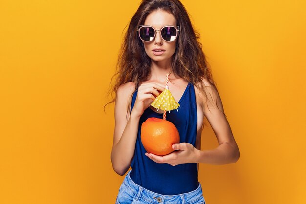 Attractive young lady in summer outfit holding fresh juicy
citrus with straw and umbrella and looking at camera while standing
on yellow