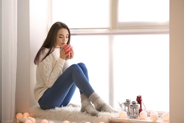 Attractive young lady with cup sitting on window sill at home