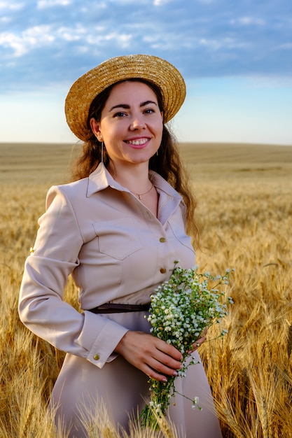 Attractive young long-haired woman in straw hat smiling and holds a wild flower bouquet in wheat field at sunrise.