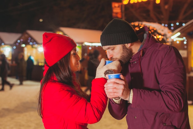 夜のクリスマスマーケットで魅力的な若い恋人たち。両手でホットカップを保持しているハンサムな男、赤い冬の服を着た素敵な女性は彼女の手でお茶で彼女の配偶者を扱います