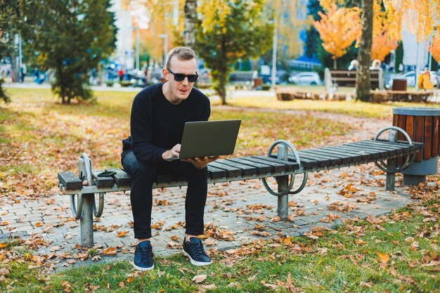 黒いセーターとサングラスをかけた魅力的な若い男性が、秋の公園のベンチに座り、ノートパソコンで作業している