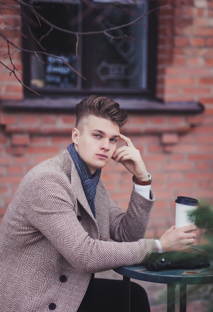 Attractive young man in coat is sitting in street cafe and drinking tea